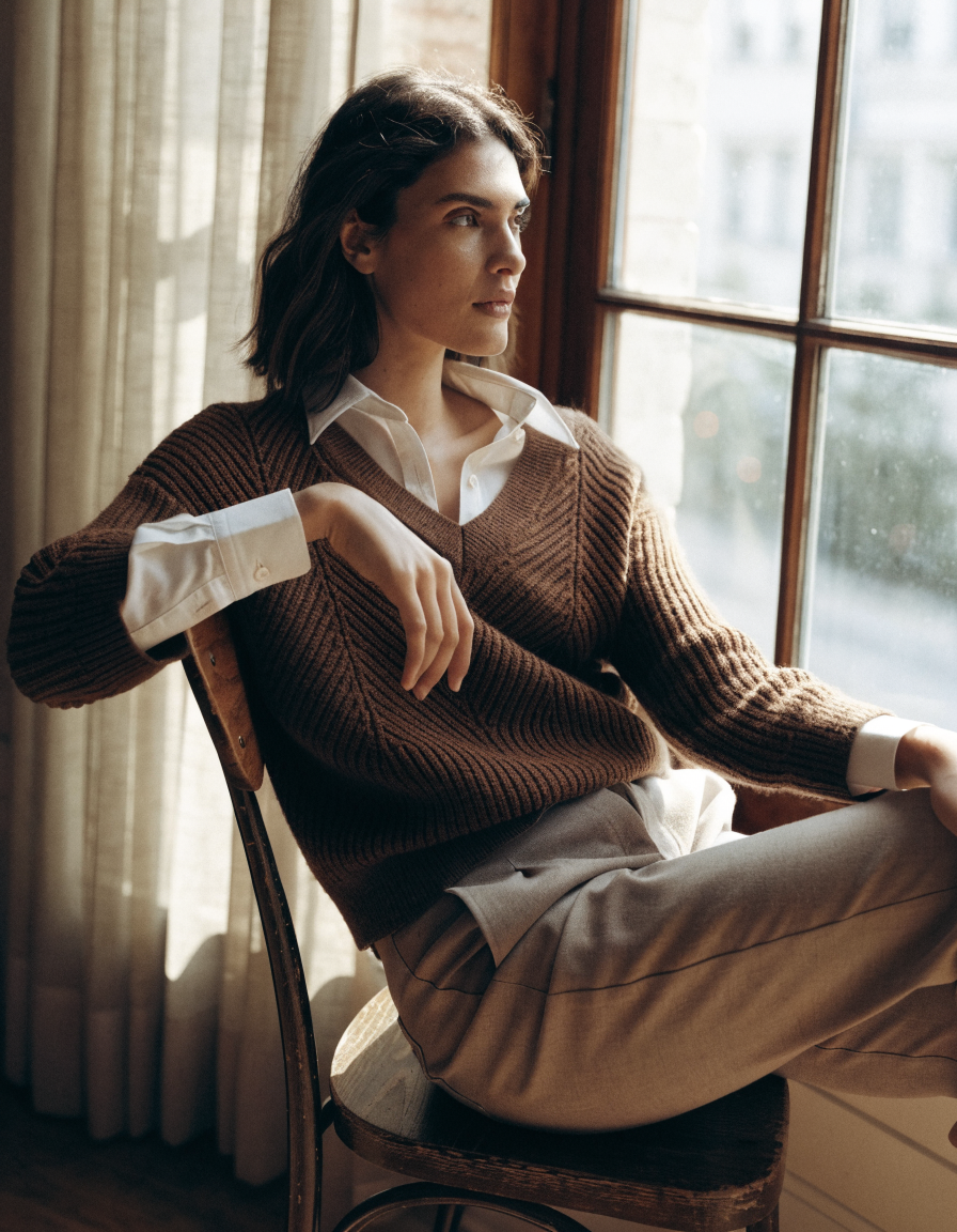 Woman wearing a brown chevron ribbed V-neck sweater over a collared shirt by a window