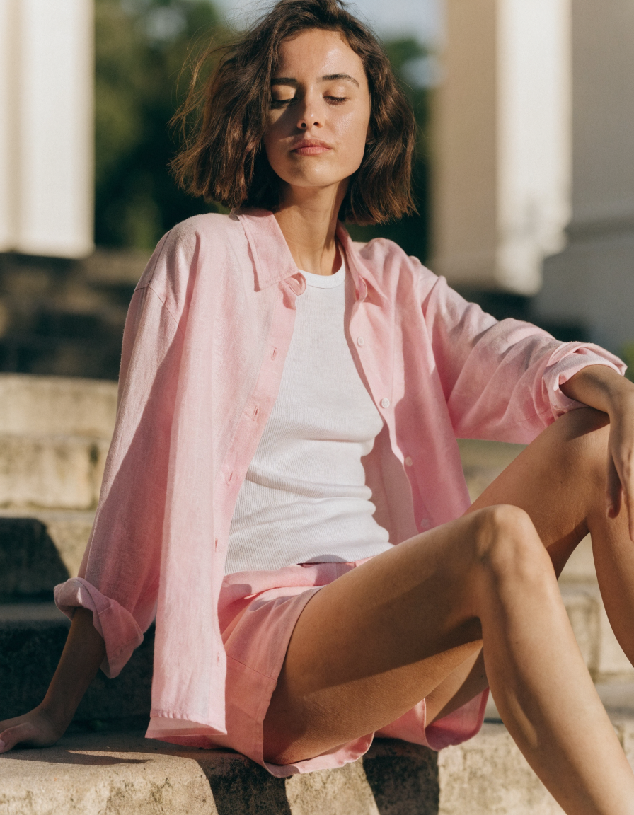 Pink woven shirt worn open over a white tank with matching shorts, seated on outdoor steps