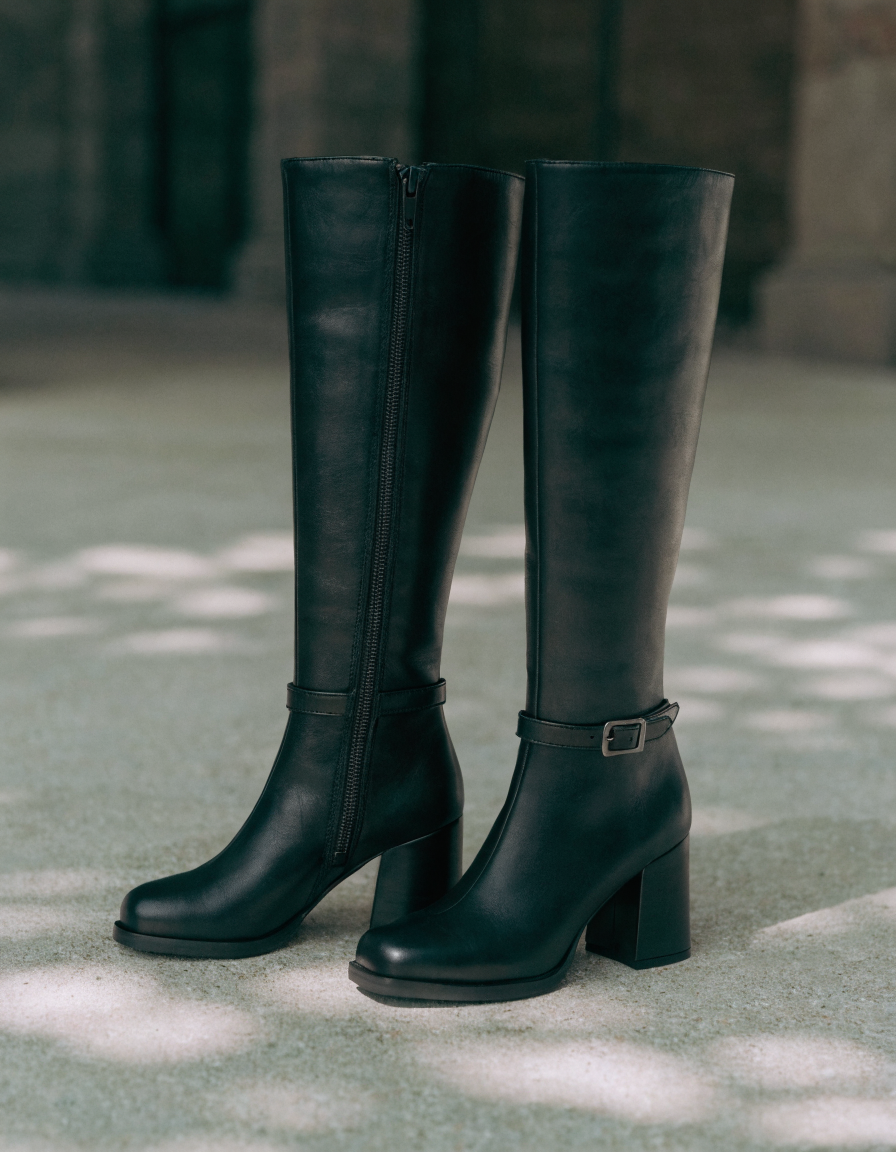 Black knee-high boots with side zip and buckle strap on pale concrete